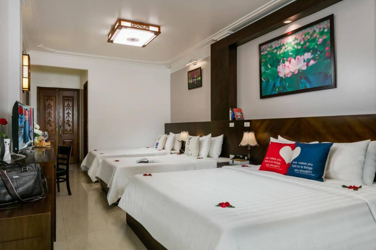 Quarto de hotel com três camas, uma escrivaninha com cadeira, uma luminária e um quadro com flores na parede.