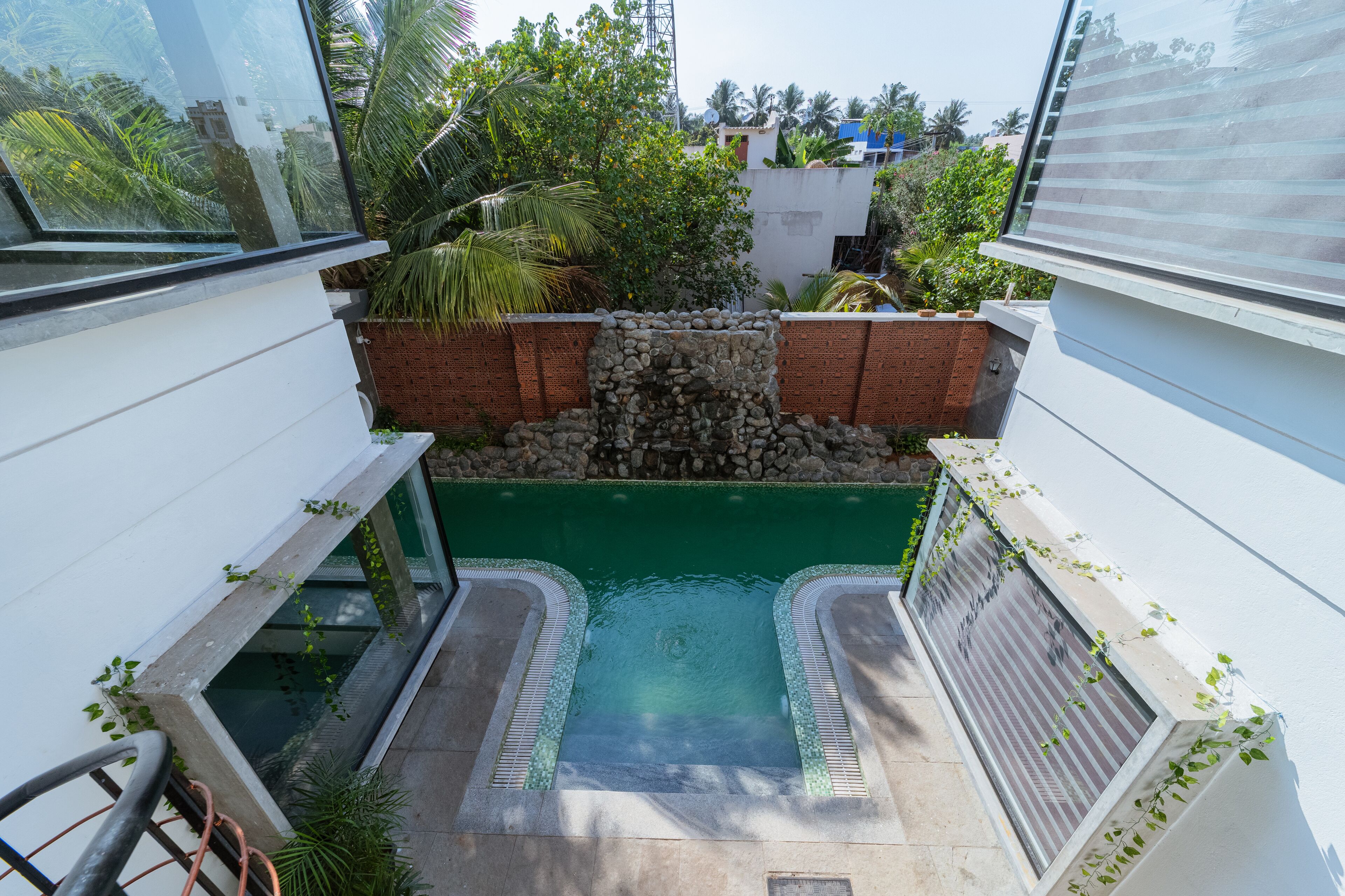A small, enclosed swimming pool with a stone waterfall feature, surrounded by lush greenery and white walls.