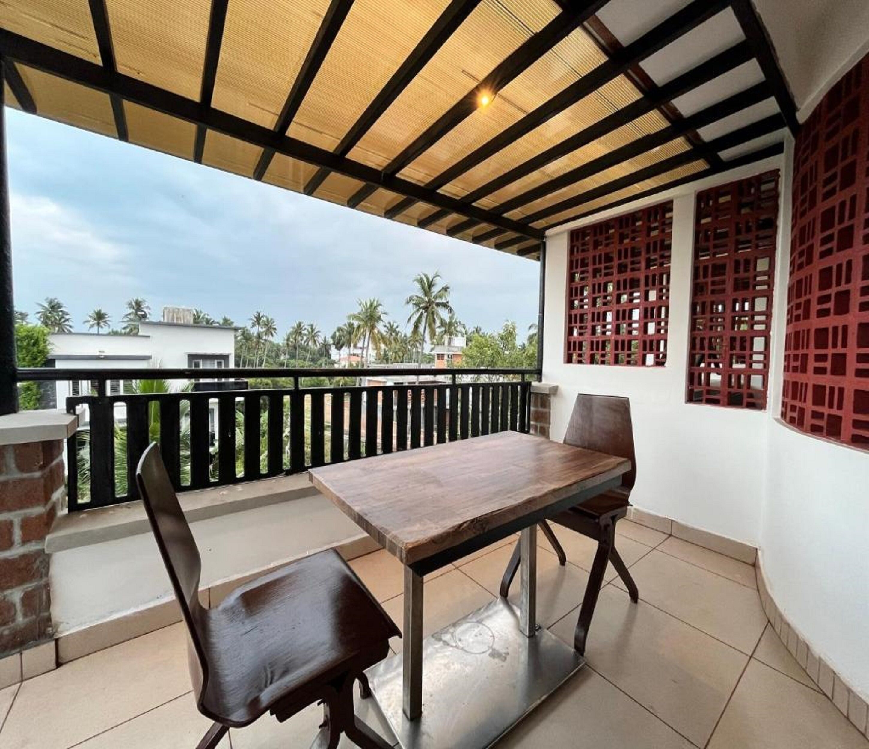 A covered outdoor seating area with a table and chairs, a view of palm trees and buildings in the background, and a red lattice window design.
