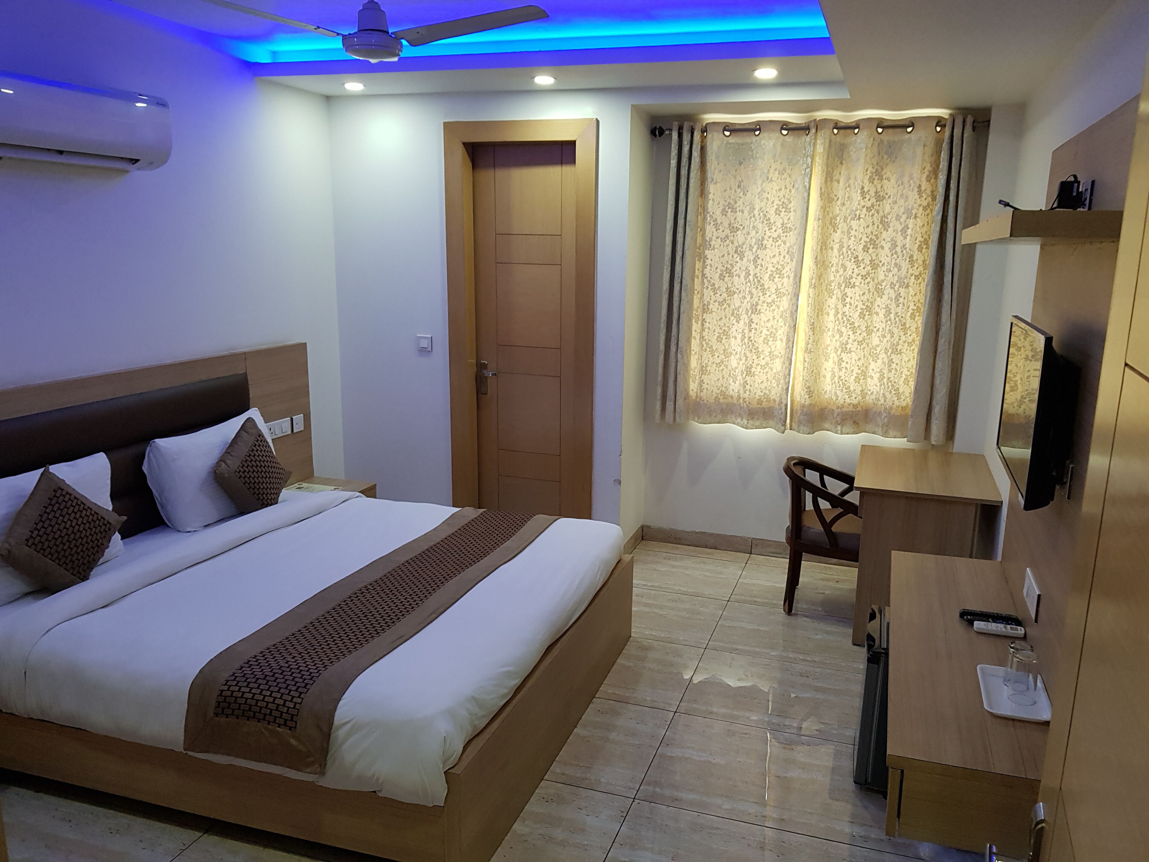 Quarto de hotel com cama, escrivaninha, cadeira, guarda-roupa, ventilador de teto e janela com cortinas.