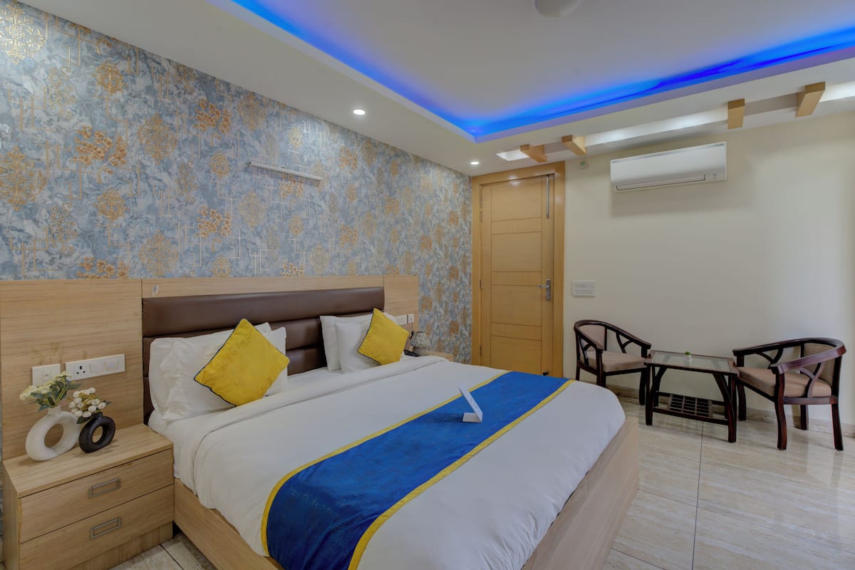 A hotel room with a large bed, wooden headboard, two bedside tables, a desk with chairs, and an air conditioning unit.