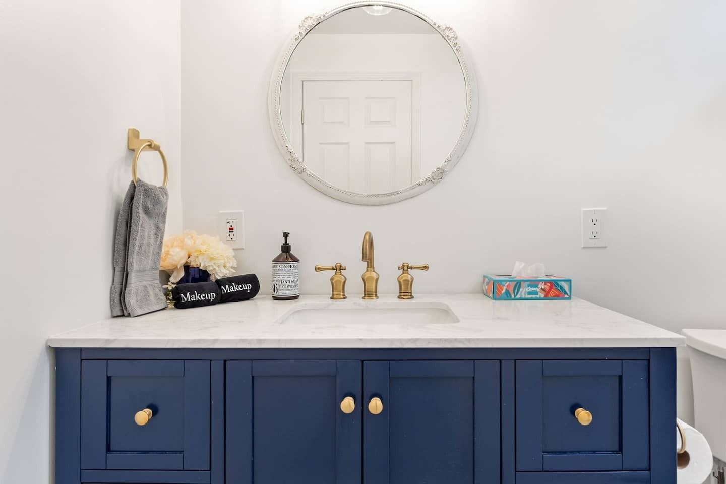 A bathroom with a white sink, gold faucets, a blue vanity with gold handles, a round mirror, and a towel rack with a gray towel.