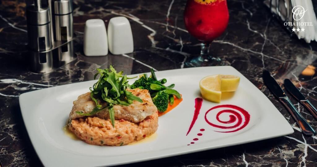 A white plate with a fish dish, greens, and lemon on a marble table.