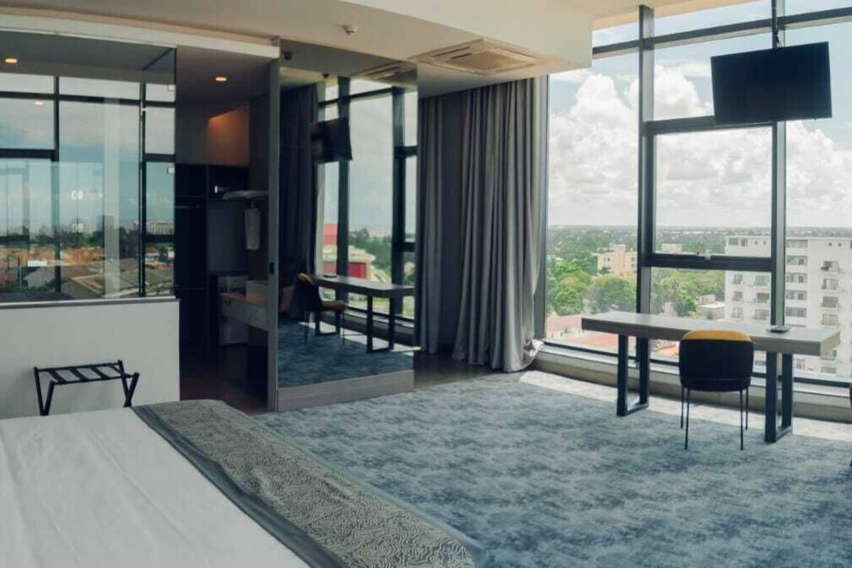 A hotel room with a large bed, a desk, a chair, and a view of the city through large windows.