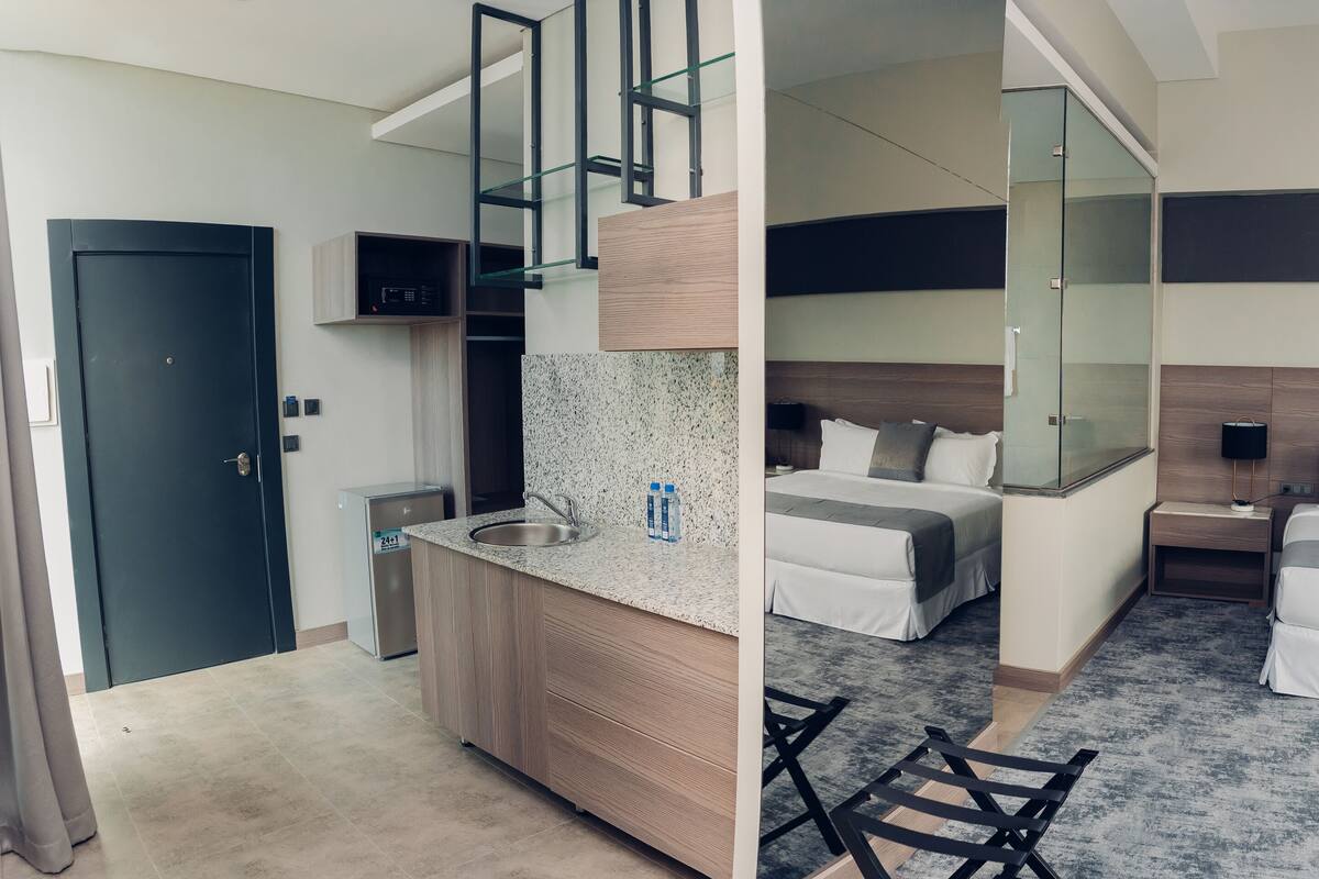 A modern hotel room with a kitchenette, two beds, a glass shower enclosure, and a large mirror.