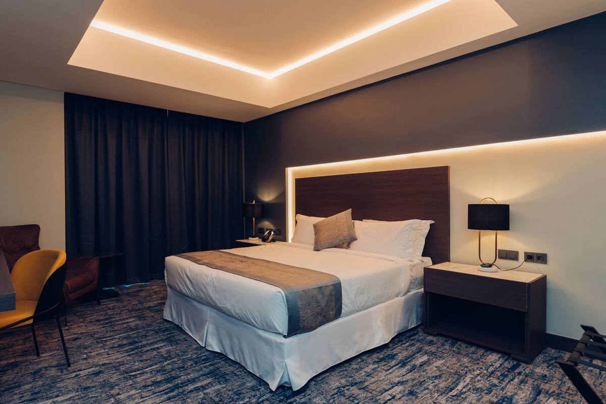 A modern hotel room with a large bed, a nightstand, a lamp, a chair, and a carpeted floor.