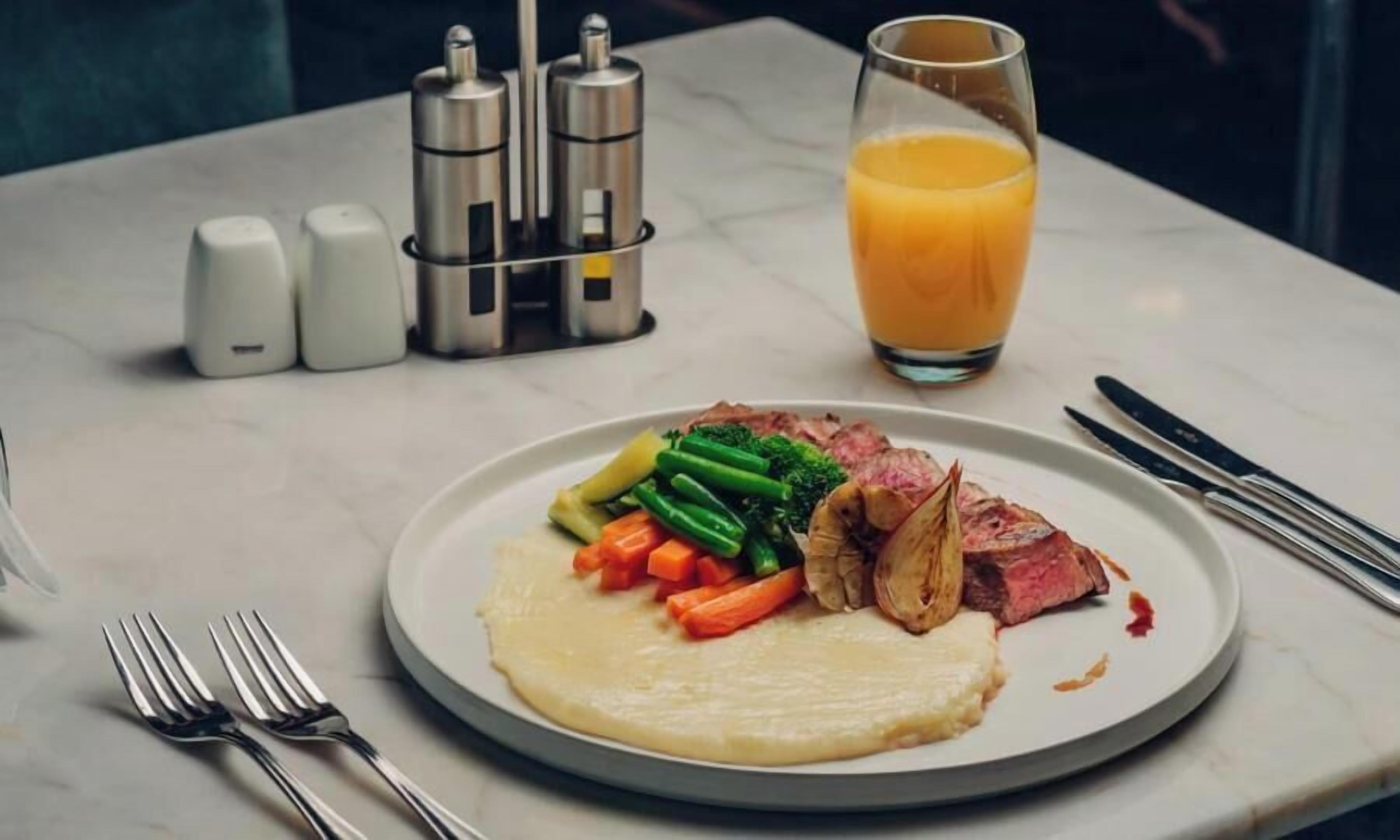 A plate of food with steak, vegetables, and a glass of orange juice on a table.