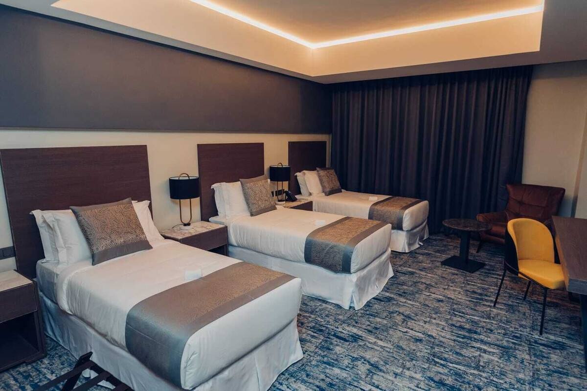 A hotel room with two single beds, a small table, a chair, and a large window with curtains.