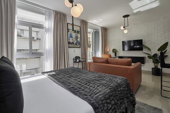 A modern hotel room with a large bed, a sofa, a TV mounted on a brick wall, and a balcony with curtains.
