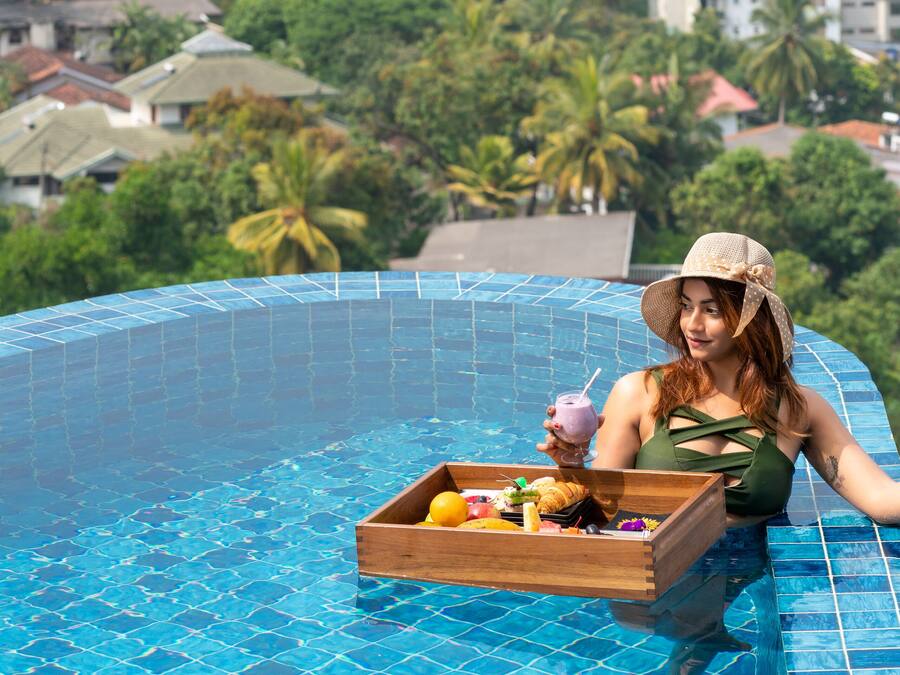 A person relaxing in a swimming pool with a tray of food and drinks, surrounded by a view of a residential area and greenery.