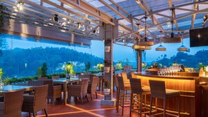 An outdoor dining area with wicker chairs and tables, a bar with stools, and a view of a forested hillside.