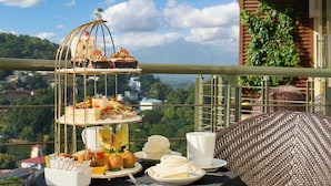 A balcony with a tiered dessert stand, plates of food, and a cup on a table with a view of a mountainous landscape.