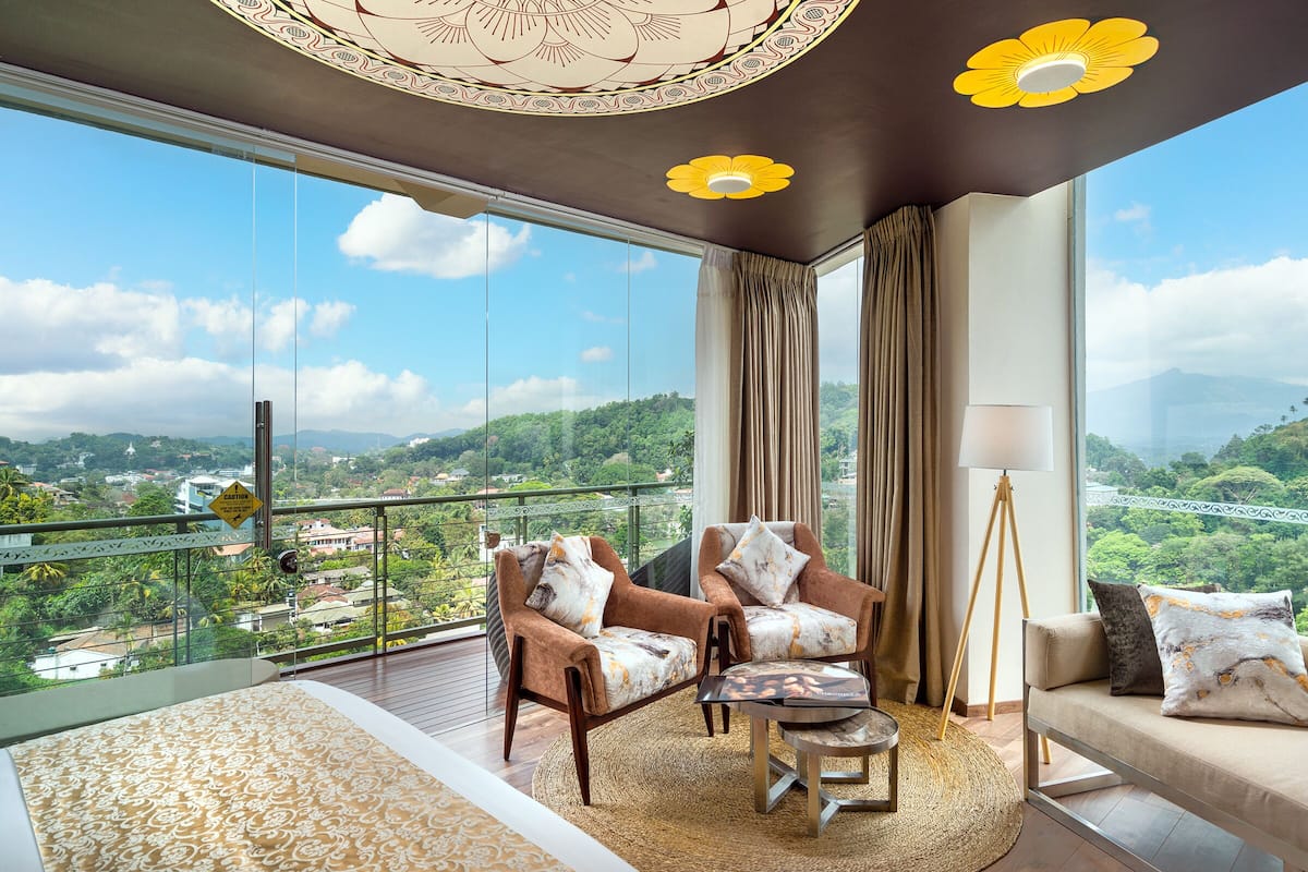 A balcony with a view of a city and mountains, furnished with two armchairs, a sofa, and a floor lamp.
