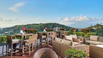 An outdoor patio with wicker furniture, tables, and chairs, overlooking a lush green landscape and a mountain in the distance.