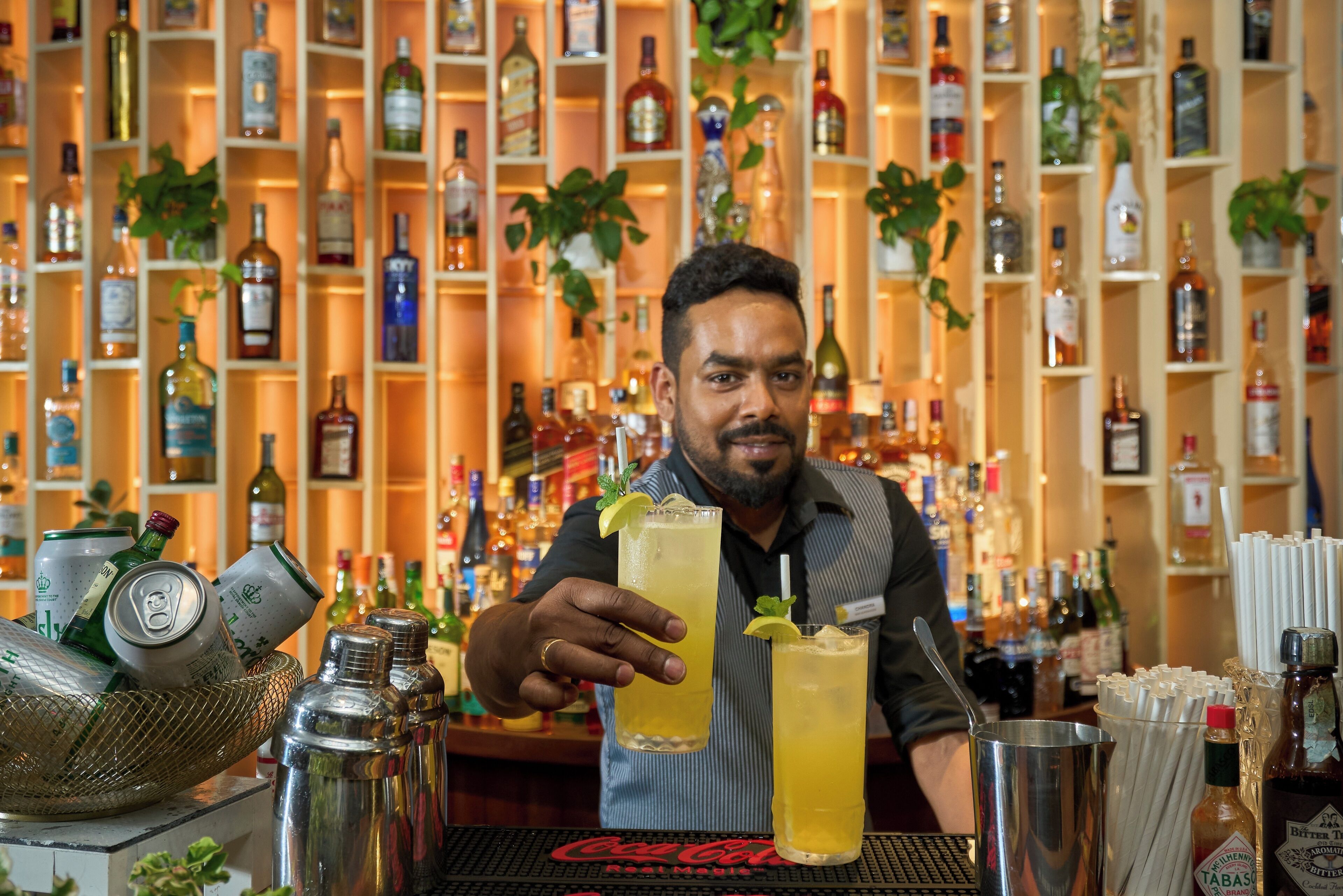 Un barman tenant deux verres derrière un comptoir, avec des étagères remplies de bouteilles d’alcool en arrière-plan.