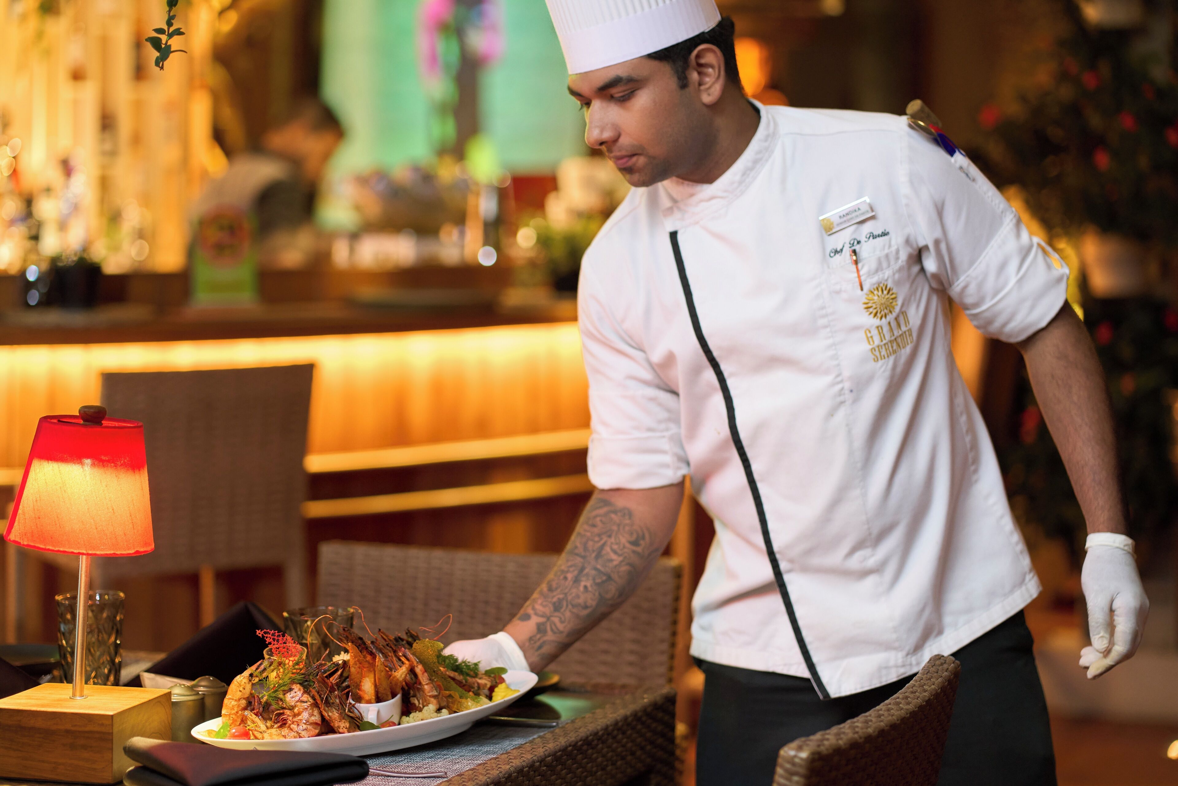 Een ober in een witte uniform met koksmuts serveert een bord eten in een restaurant met een rode lamp op tafel.