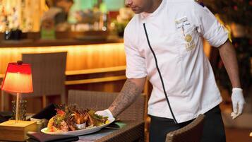 Um garçom de uniforme branco e chapéu de chef servindo um prato de comida em um restaurante com uma luminária vermelha sobre a mesa.