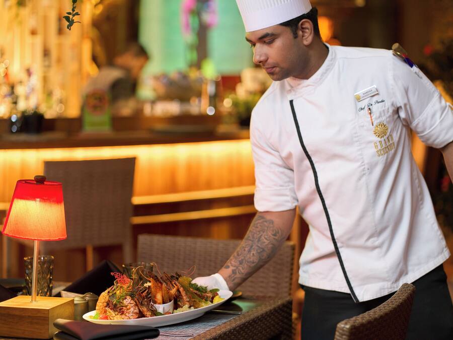 Um garçom de uniforme branco e chapéu de chef servindo um prato de comida em um restaurante com uma luminária vermelha sobre a mesa.