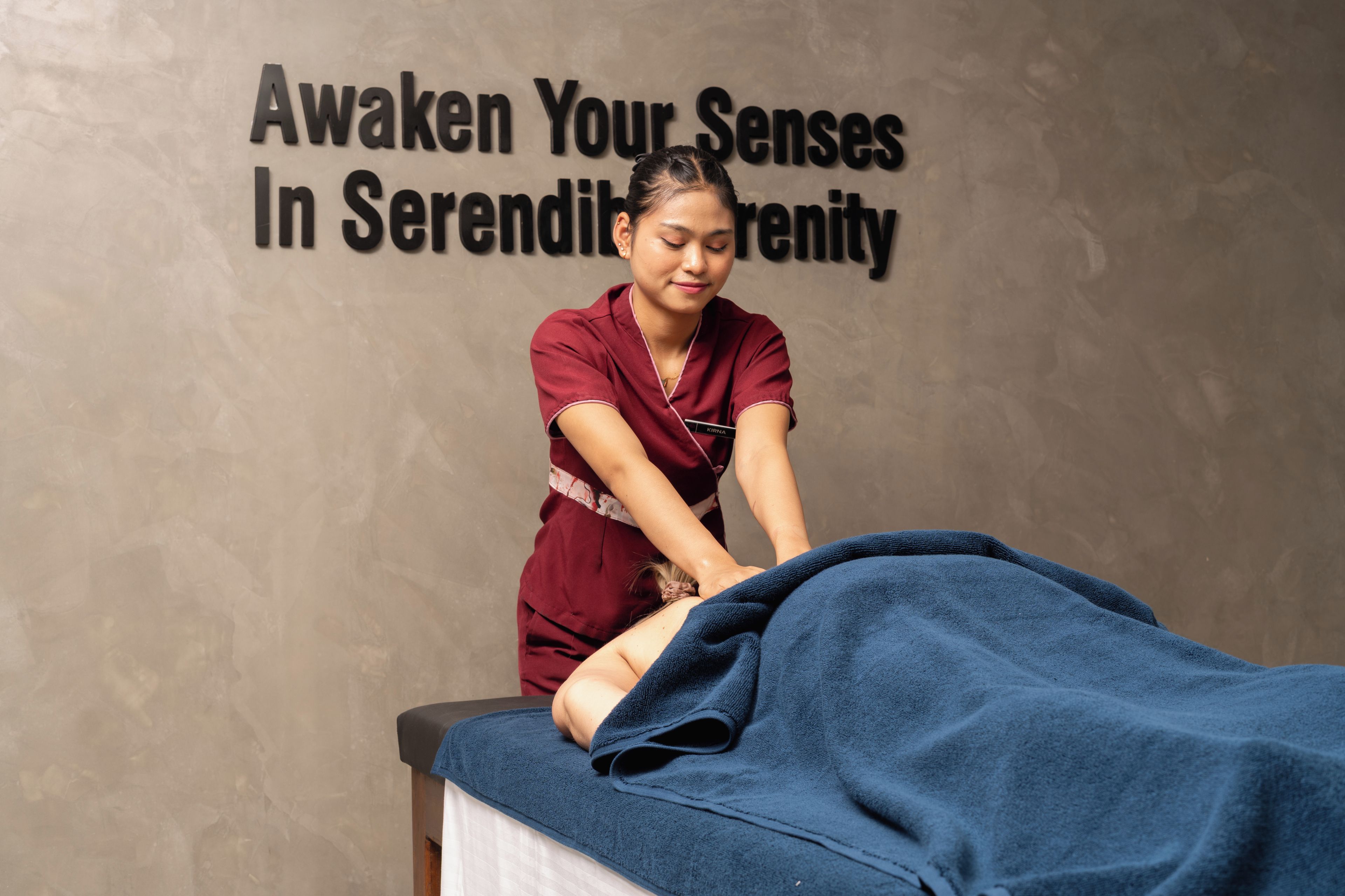 A massage therapy session with a person lying on a treatment table covered with a blue towel, and a wall with text that reads 'Awaken Your Senses In Serenity'.