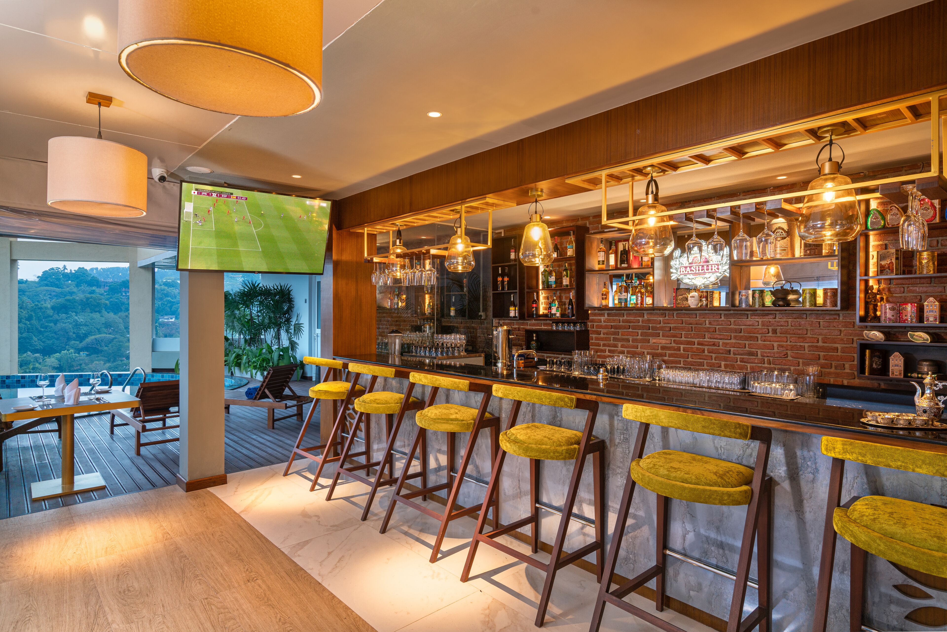 A bar area with wooden bar stools, a TV screen displaying a sports game, and a view of the outdoors.
