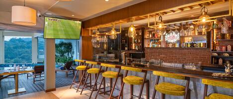 A bar area with wooden bar stools, a TV screen displaying a sports game, and a view of the outdoors.