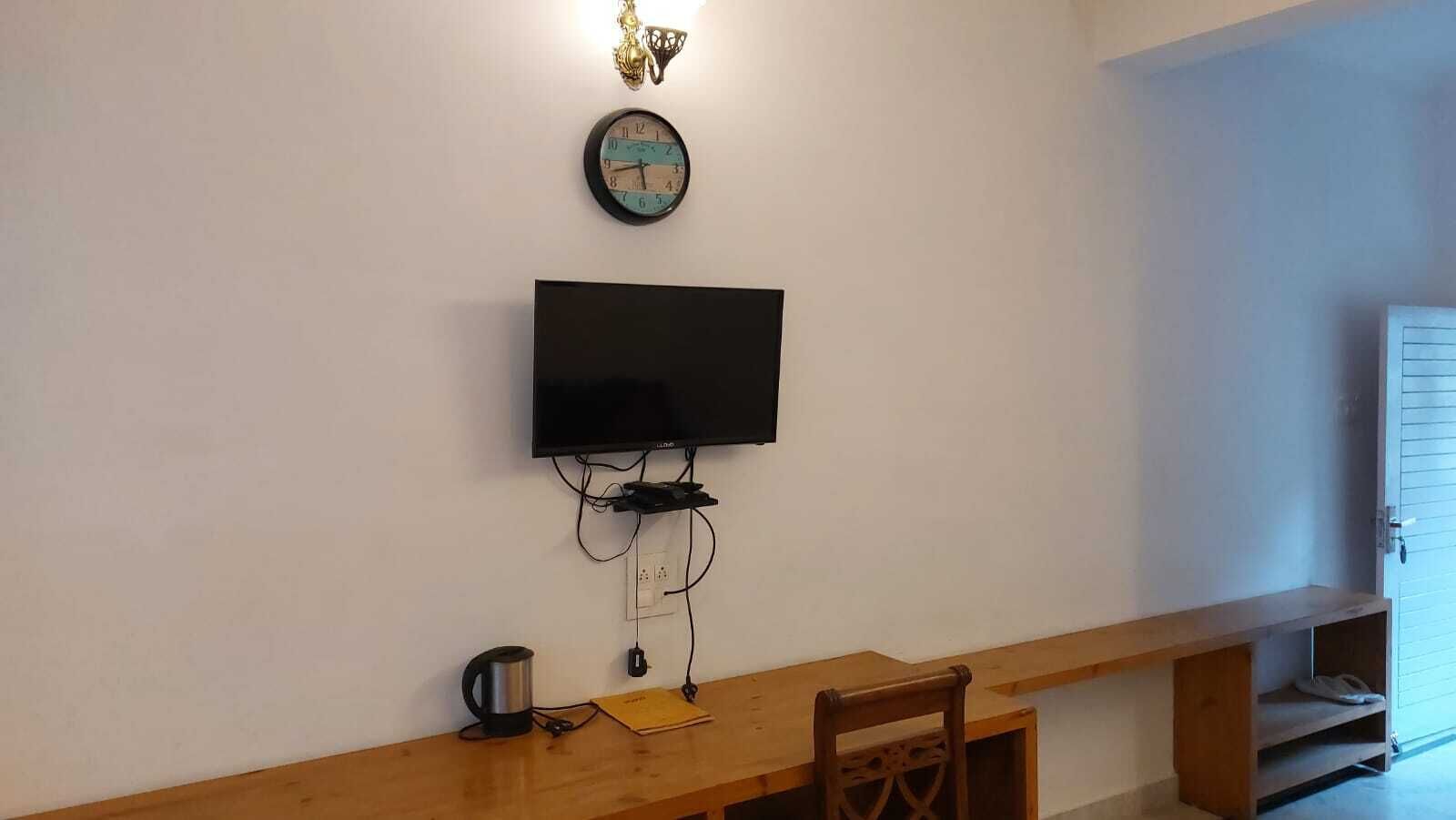 Una habitación con una televisión de pantalla plana montada en la pared, un banco de madera, una mesita con un hervidor y una puerta con ventana de vidrio corredera.