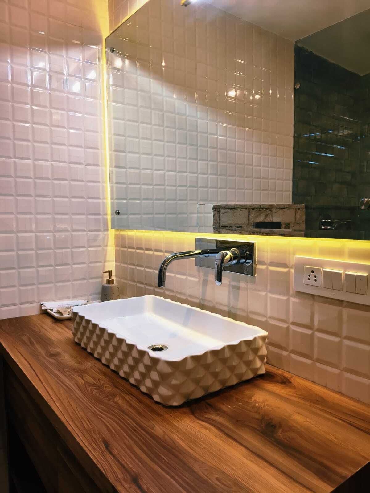 A bathroom with a white tiled wall, a wooden vanity, a white sink with a geometric pattern, a mirror, and a wall-mounted light fixture.