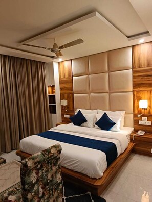 Une chambre d’hôtel avec un grand lit, un ventilateur de plafond, une tête de lit en bois et un fauteuil à motifs floraux.