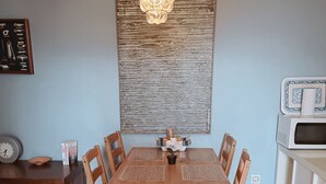 Et spiseområde med et træbord, stole, en hængende lampe, et billede på væggen og en mikrobølgeovn i et hvidt skab.