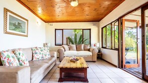 A living room with a wooden ceiling, a beige sofa with floral cushions, a wooden coffee table, and large windows offering a view of greenery. - Hilltop Hideaway (eMdloti)