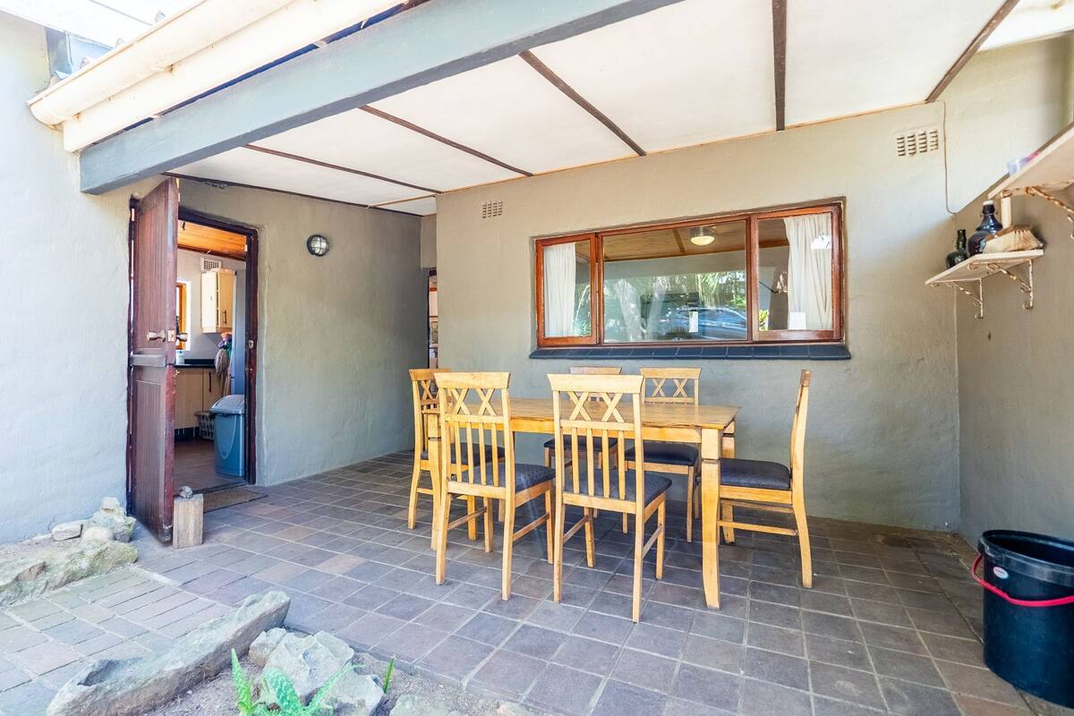 A covered patio area with a wooden table and chairs, a window with curtains, and a black trash bin.