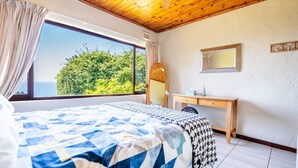 A bedroom with a large bed, a wooden desk, and a view of greenery through a window. - Hilltop Hideaway (eMdloti)