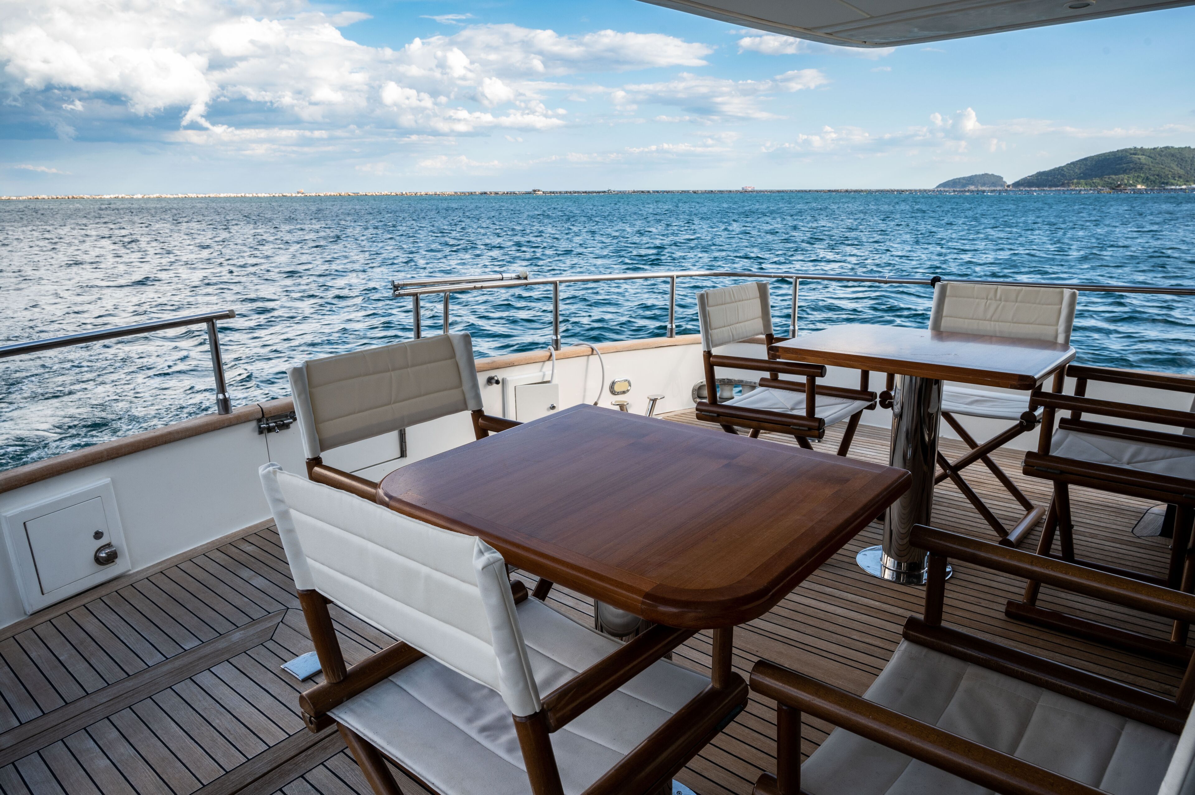 Cubierta de barco con mesa de madera y dos sillas acolchadas, barandilla y vista al mar y la costa lejana.