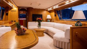 A yacht interior with a round dining table, a sofa, a small side table, and a large window with a view of the sea.
