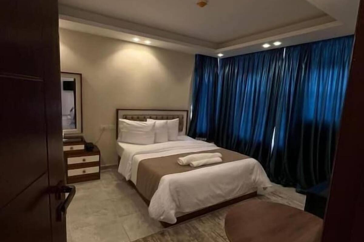 A hotel room with a bed, a bedside table, a chair, and a large window with curtains.