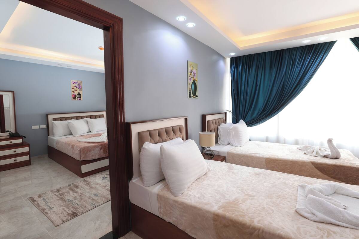 A hotel room with two beds, a large mirror, a nightstand, a lamp, and a window with curtains.