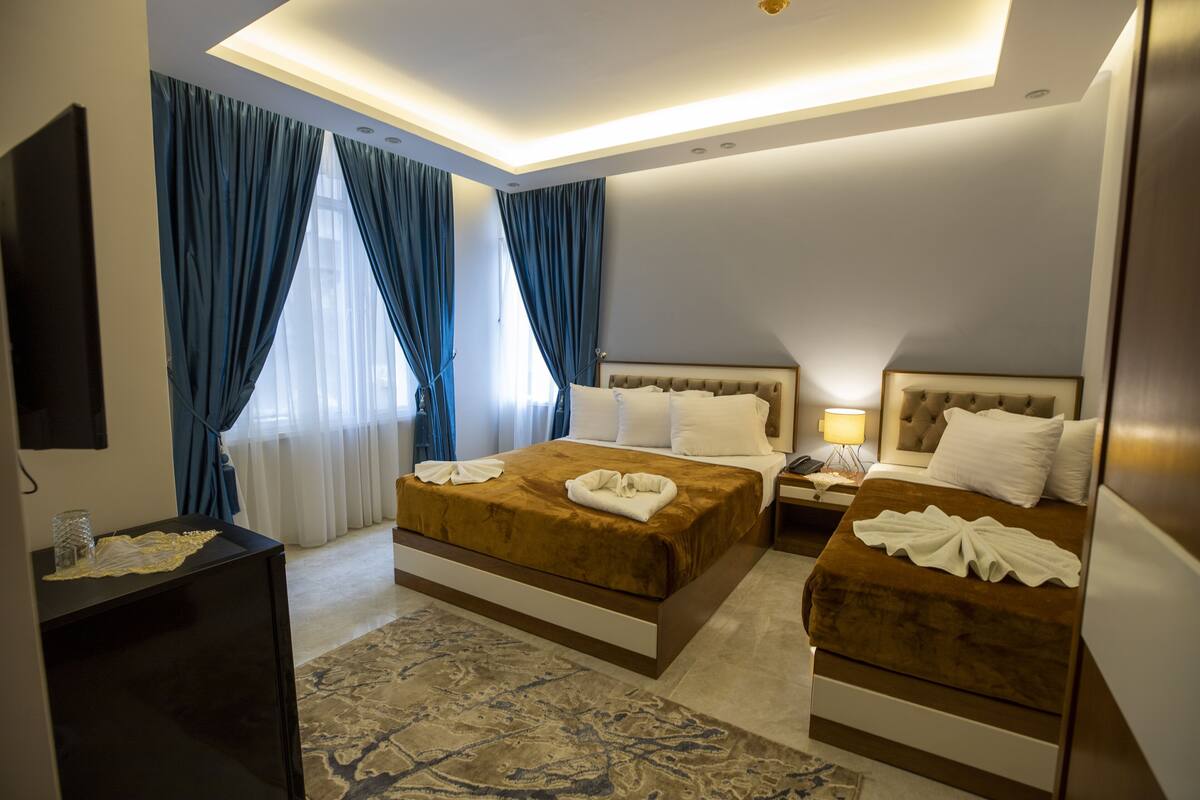 A hotel room with two beds, a flat-screen TV, a desk, and a large window with curtains.