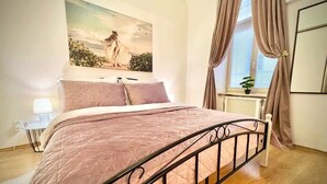 A bedroom with a bed, a nightstand, a lamp, a window with curtains, a radiator, and a painting on the wall. - Elegant Apartment on Hlavná Košice (Košický kraj)