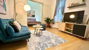 A living room with a blue sofa, a small round table, a TV on a wooden stand, and a large window with curtains. - Elegant Apartment on Hlavná Košice (Košický kraj)