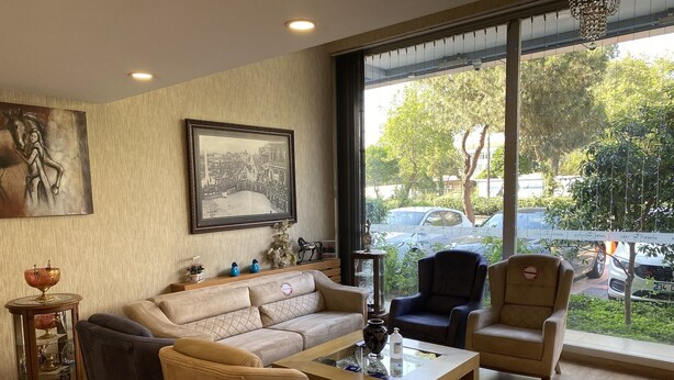 A modern living room with a beige sofa, two armchairs, a coffee table, and a large window offering a view of parked cars and greenery outside. - Aydın Park Otel (Aydin)