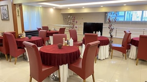 A hotel dining area with round tables covered in red tablecloths, red upholstered chairs, and a large flat-screen TV mounted on the wall. - Aydın Park Otel (Aydin)