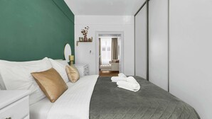 A neatly made bed with a grey quilt, white pillows, and a brown accent pillow. There is a white bedside table with a lamp and a vase with dried flowers. A white wardrobe is visible in the background. - Trendy Apartment in Centrum 1 (Bratislavský kraj)