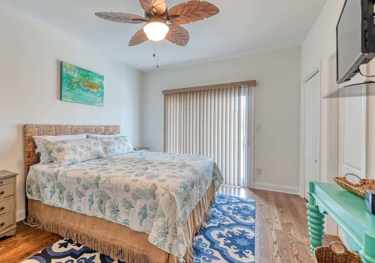 Une chambre à coucher avec un lit, un ventilateur de plafond, une fenêtre avec des stores, un plancher en bois et une table de chevet verte.