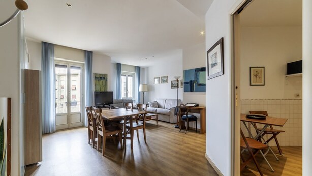 A modern living room with a sofa, TV, and dining area with a table and chairs. - GuestHost - Cozy apartment of 90 sqm, can accommodate up to 5 people, located on the sixth floor of a building with a lift (NOT suitable for disabled people).The property is located in an area well connected by public transport, the metro line B Piramid (Roma)