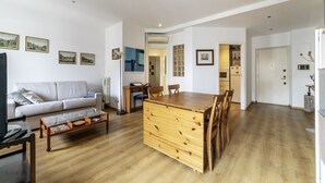 A living room with a sofa, coffee table, wooden dining table, and chairs. There is a kitchen area with cabinets and a refrigerator. - GuestHost - Cozy apartment of 90 sqm, can accommodate up to 5 people, located on the sixth floor of a building with a lift (NOT suitable for disabled people).The property is located in an area well connected by public transport, the metro line B Piramid (Roma)