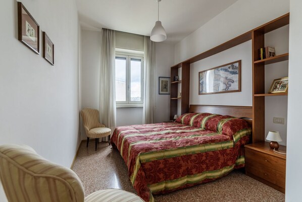 A bedroom with a bed, a chair, a desk, a shelf with books, and a window with curtains.