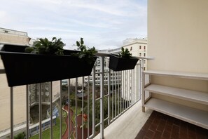 A balcony with black planters containing green plants, a view of a courtyard with buildings, and a white railing.