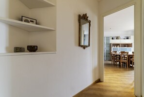 A hallway with a wooden floor, a white wall with a mirror, and a dining area in the background.