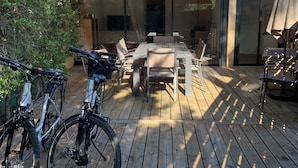A wooden deck with outdoor furniture, bicycles, and a covered patio area. - Chalet in a shady residence with swimming pool (Occitanie)
