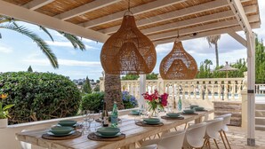 An outdoor dining area with a wooden table set for six, surrounded by white chairs, under a pergola with hanging wicker lamps. - Large Villa with sea view in delightful Benissa Montemar (Costa Blanca North) (Benissa)